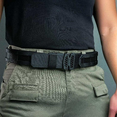 Person wearing a tactical belt with a black buckle on green pants against a gray background