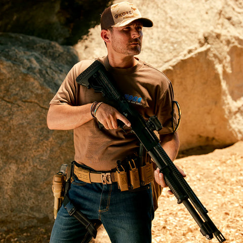 Man holding a rifle in a rocky outdoor environment, wearing tactical gear and carrying a black tactical pouch with multiple attachment loops and adjustable straps.