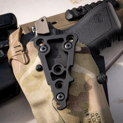 Black tactical mount on a camouflage surface with a gun in the background